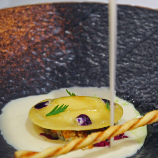 White sauce being served over a bread twirl and ravioli pasta in a charcoal stone bowl