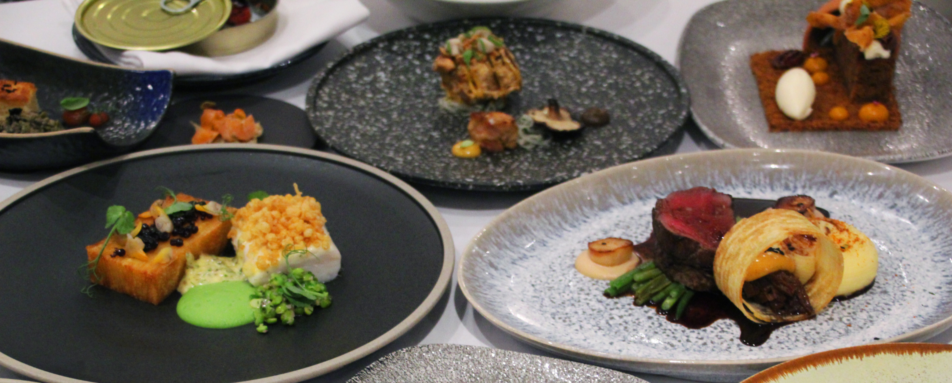 A table full of fine dining plated food on a variety of different coloured plates