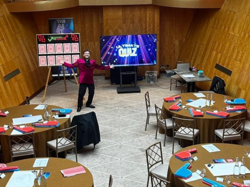 A man in red a suit infront of a TV and four round tables set-up for a quiz