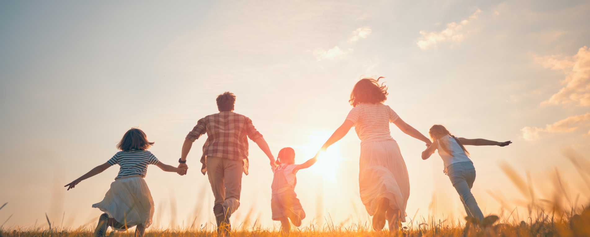 A family of five running through a grass field away from the camera, towards the sunset