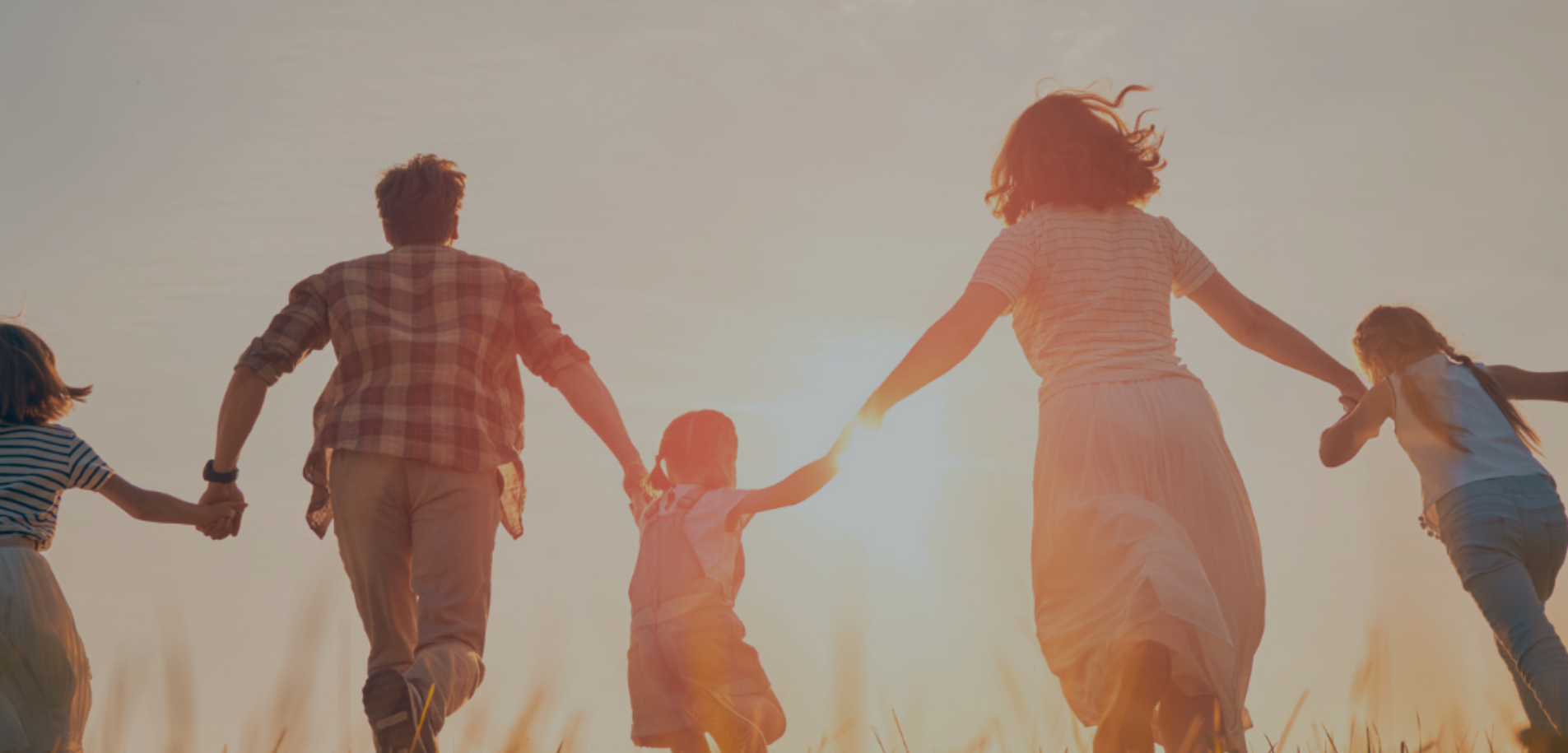 A family of five running through a grass field away from the camera, towards the sunset