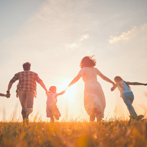 A family of five running through a grass field away from the camera, towards the sunset