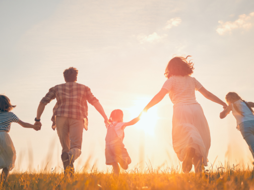 A family of five running through a grass field away from the camera, towards the sunset
