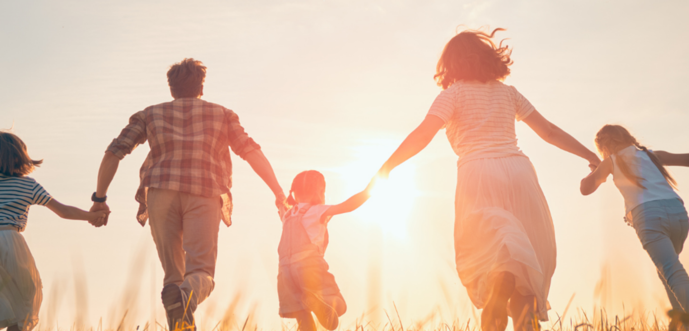A family of five running through a grass field away from the camera, towards the sunset