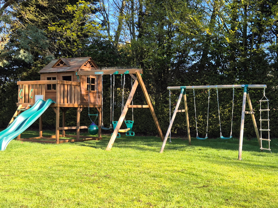 A wooden playground with slide and swing in a grass area