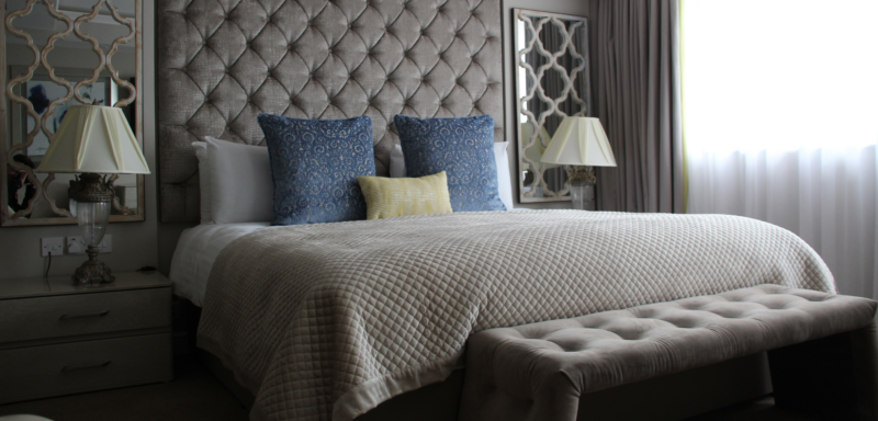 A luxurious looking king size bed with grey duvet, blue cushions and a big grey headboard. With an ottoman cushion at the end of the bed.