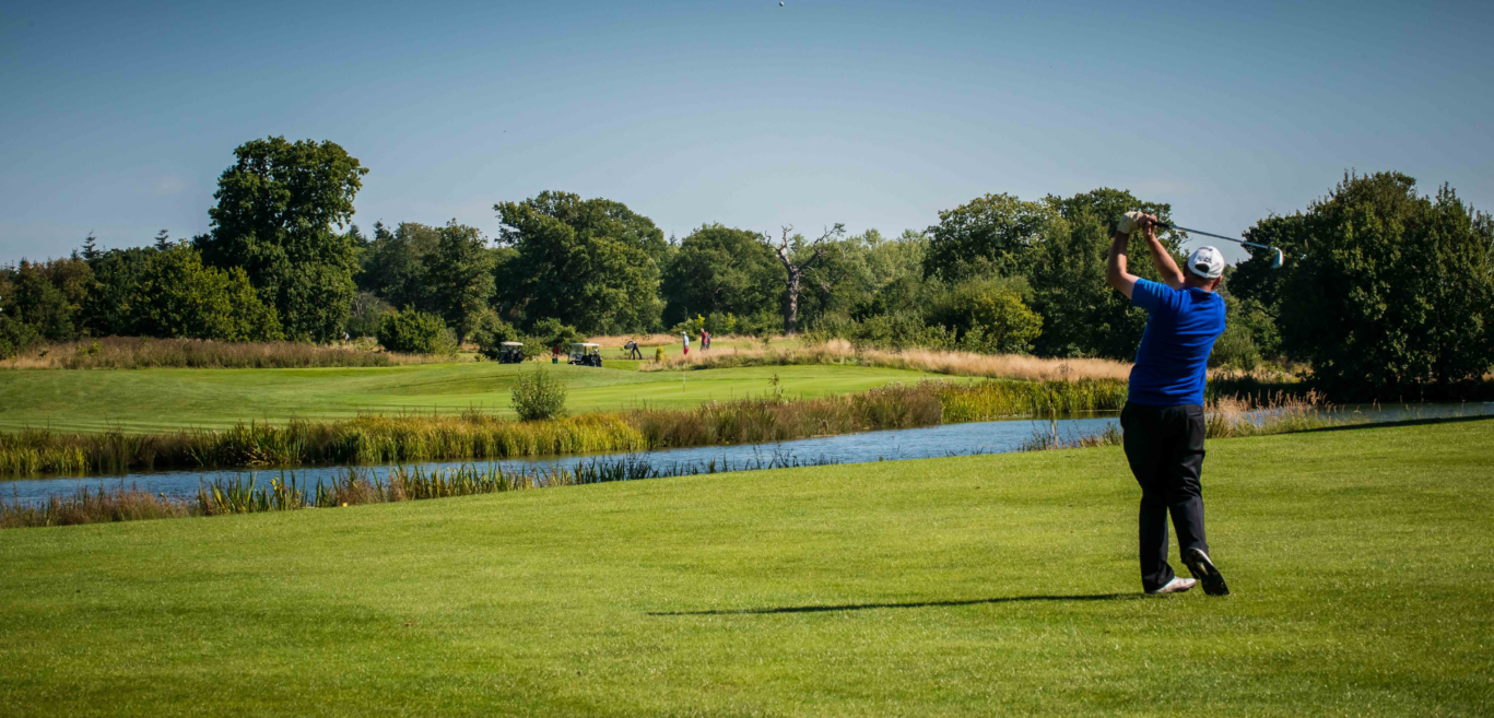 A golfer mid-swing facing down the greens on a sunny day, with a lake right next to him