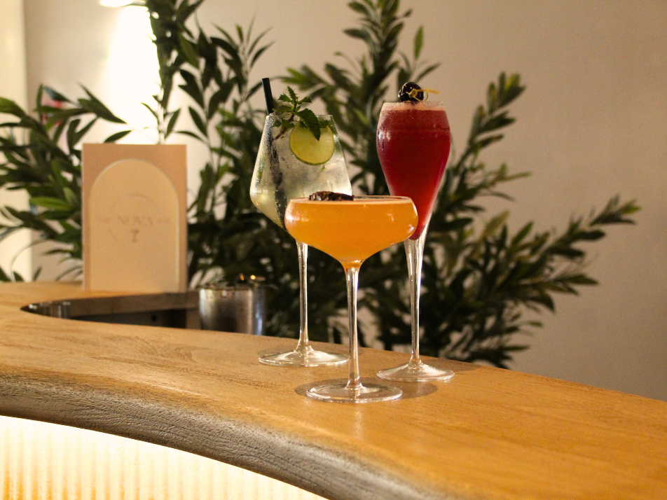 A collection of colourful cocktails sitting on top of a lit up wooden bar top with a green flower bush behind them