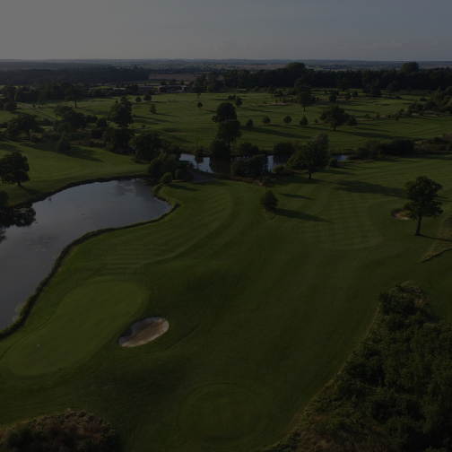 Aerial view of Whittlebury Park's green golf course with sand banks and a lake dotted in