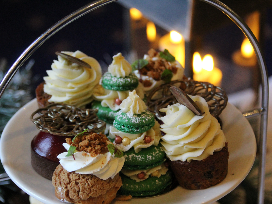 A plate of afternoon tea christmas sweet treats, with xmas tree macarons, choux buns and black forest tarts, with festive lights behind the plate