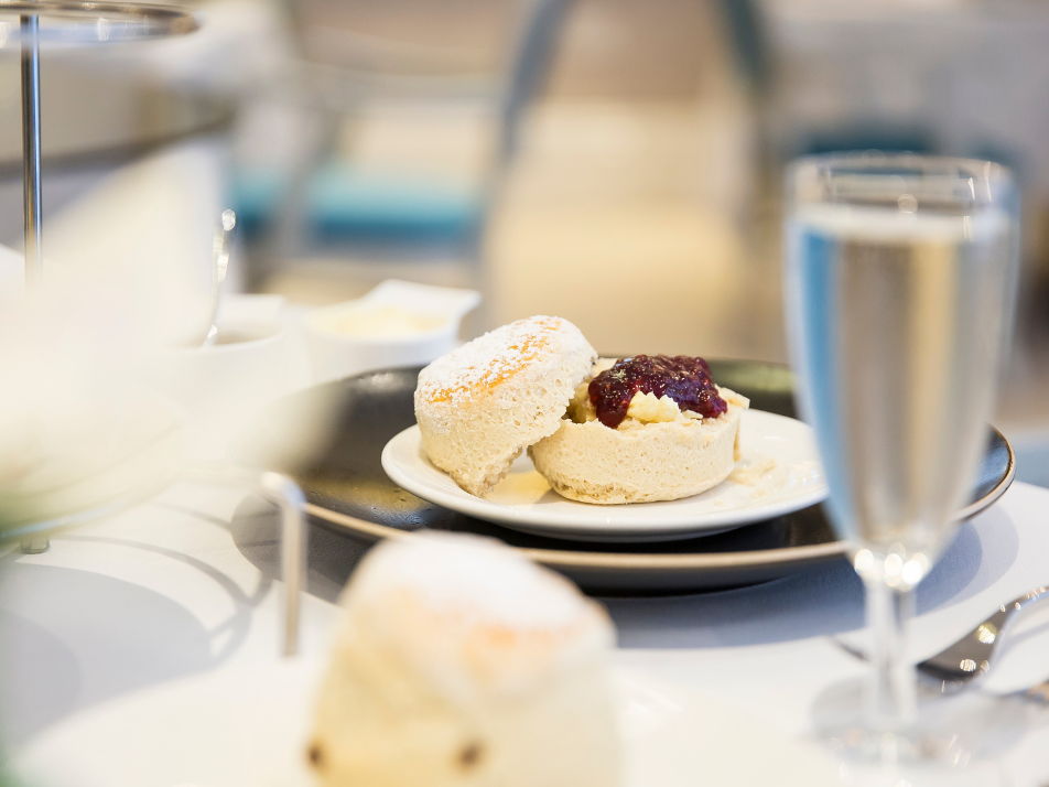 A cut in half scone with jam and cream spreaded on one side, served on a small white plate along with a glass of Prosecco