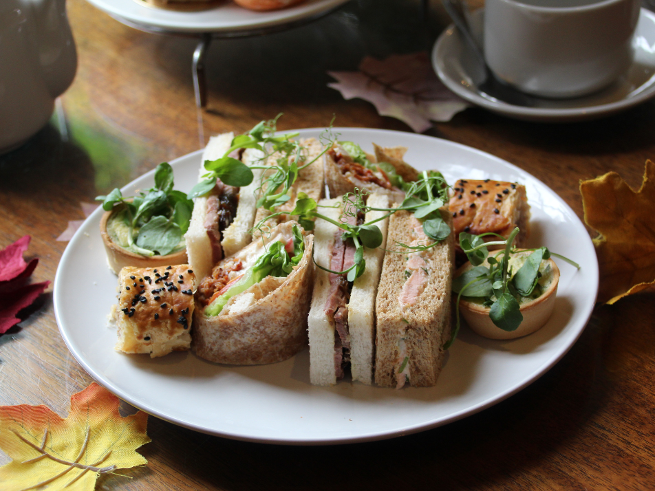 An afternoon tea plate of delicious finger sandwiches, filled wraps and sausage rolls, decorated with autumn leaves around the plate