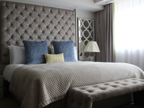 A luxurious looking king size bed with grey duvet, blue cushions and a big grey headboard. With an ottoman cushion at the end of the bed.